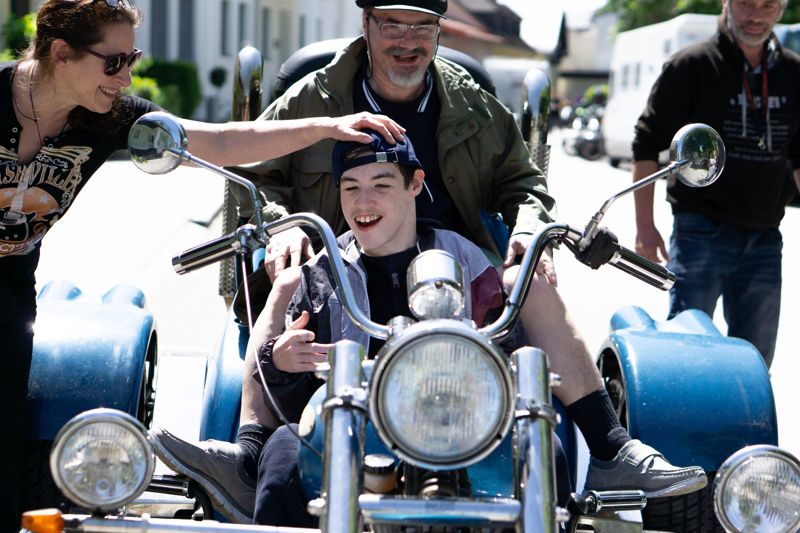 Ein junger Mann sitzt lachend auf einem Motorrad, umringt von Begleiterinnen und Begleitern.