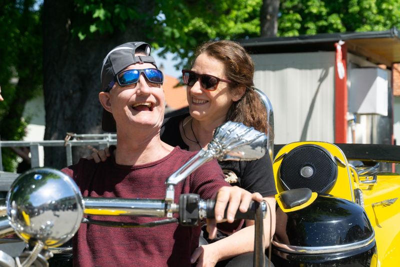 Ein Mann mit Sonnenbrille lacht auf einem Motorrad, eine Begleiterin steht lächelnd neben ihm.