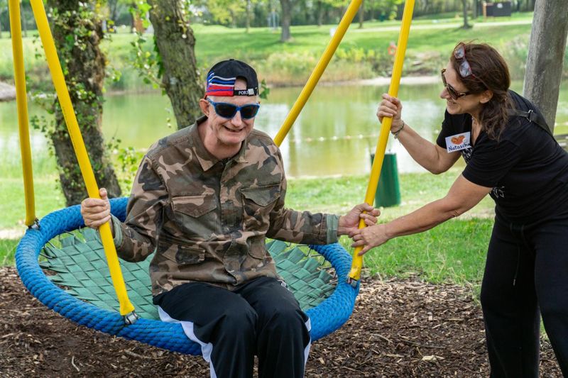 Ein Mann mit Sonnenbrille sitzt auf einer Nestschaukel am See, eine Betreuerin hält die Seile.