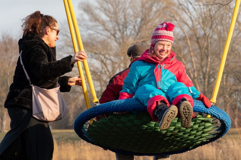 Eine junge Frau in Winterjacke liegt lachend in einer Nestschaukel, eine Begleiterin schaukelt sie an.